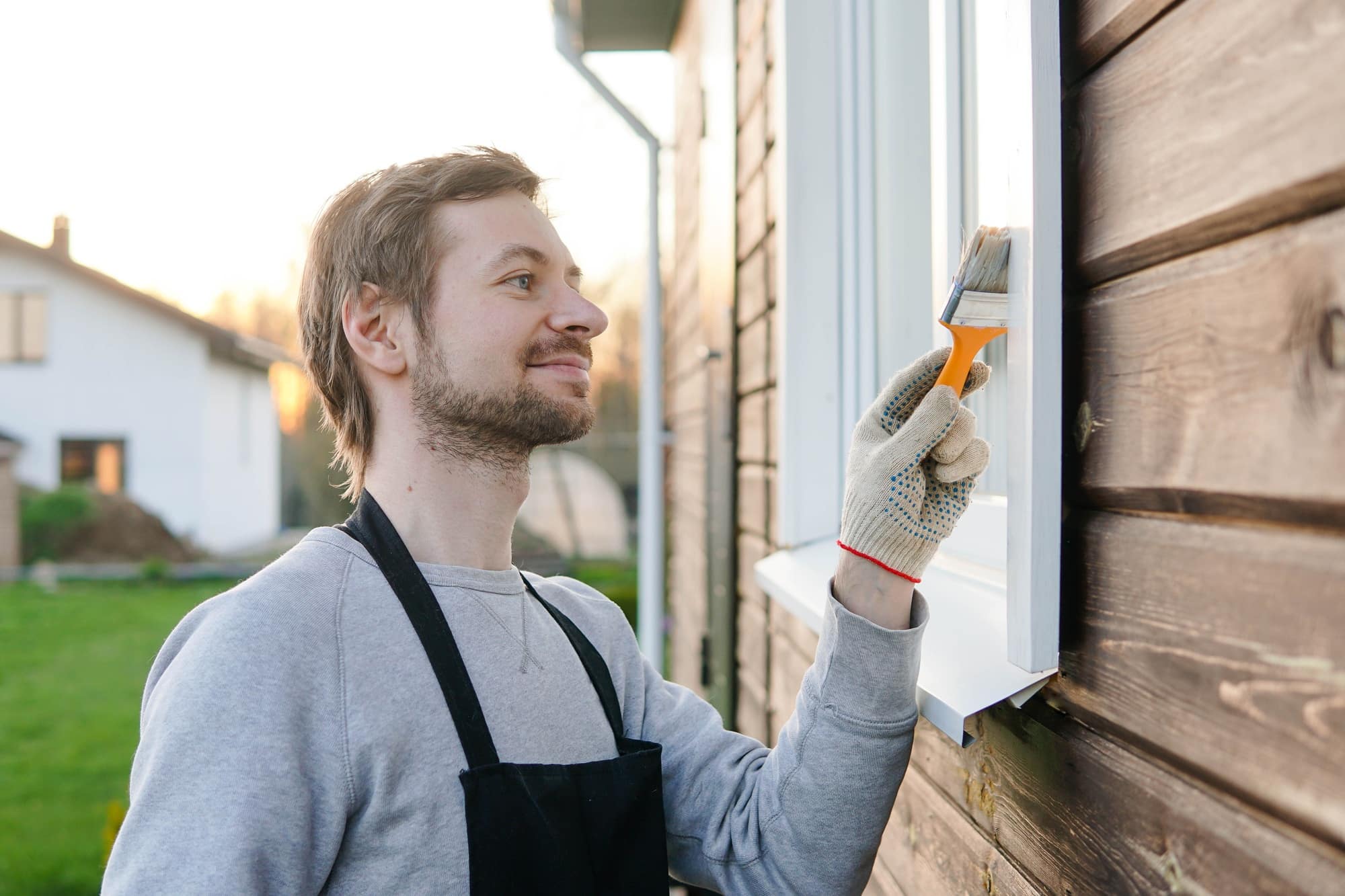 Expert Interior and Exterior Painting | King & Pierce Counties Young smiling man in apron painting window of his house with paint brush.